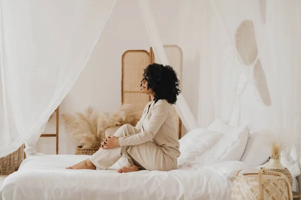 Beautiful African American young woman sitting on bed in home in white pajamas. — Stock Photo, Image