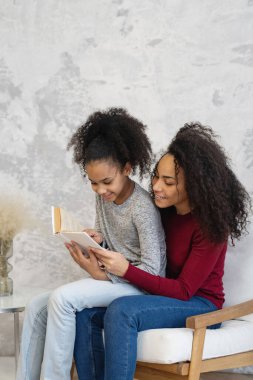 Afro-Amerikalı anne ve kızı kitap okuyorlar. Evdeki sandalyede oturuyorlar..
