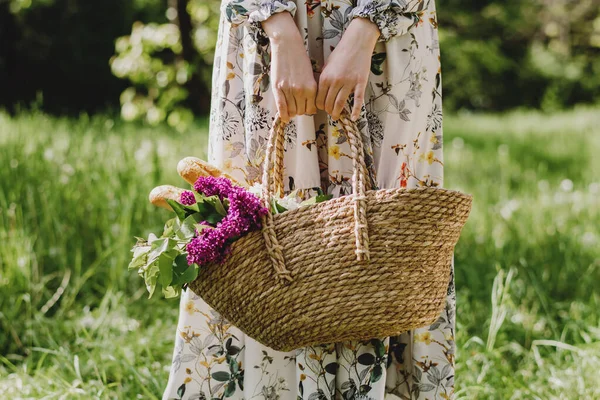 Feminino segurando cesta de piquenique com alimentos e buquê de lilases ao ar livre. — Fotografia de Stock