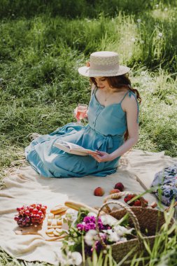 Hasır şapkalı sevimli kadın dışarıda piknikte kitap ve şarapla dinleniyor..