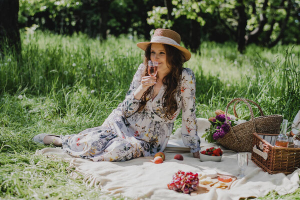 Young woman in summer dress and straw hat sitting with rose wine, cheese, fresh fruit and baguette on blanket.