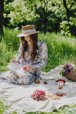 Young woman in straw hat wearing summer dress reading a book while relaxing in the park.