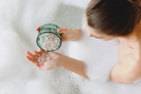 거품 목욕을 하고 손에 바다 목욕 소금을 들고 앉아 있는 여자의 맨 위 사진. — 스톡 사진