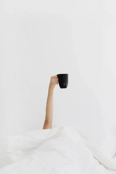Γυναικείο χέρι ξαπλωμένο στο κρεβάτι και κρατώντας κούπα με καφέ. — Φωτογραφία Αρχείου