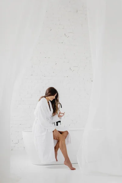 Привлекательная молодая женщина сидит на ванне и наносит крем. — стоковое фото