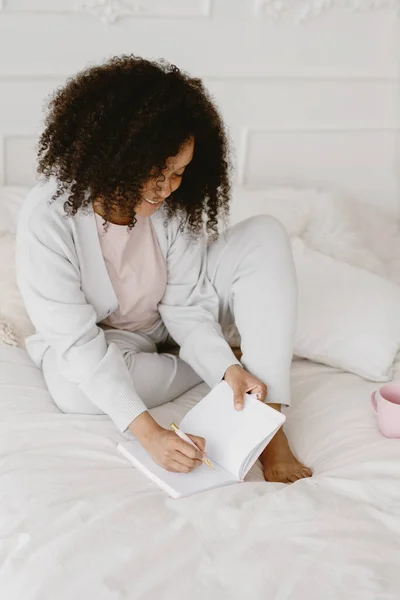 Αφροαμερικανή που κάθεται στο κρεβάτι και κρατάει σημειώσεις στο ημερολόγιο.. — Φωτογραφία Αρχείου
