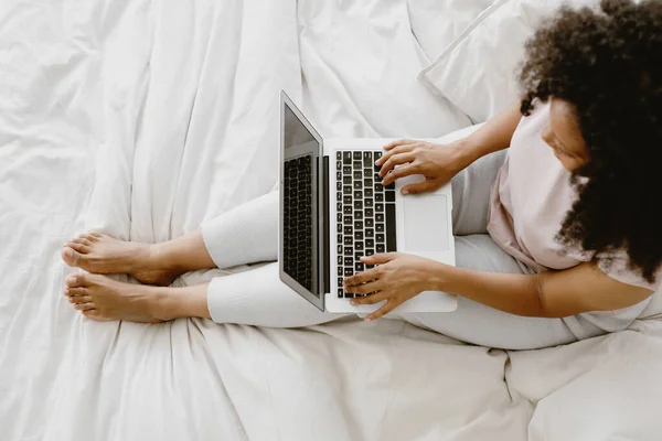 Jonge schattige Afro-Amerikaanse vrouw zitten op het bed en typen op laptop. — Stockfoto