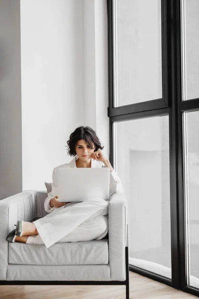 Jonge zakenvrouw zit op stoel met laptop bij het raam. — Stockfoto