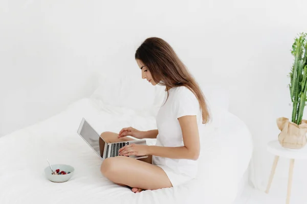 Jonge vrouw eet ontbijtgranen op bed in de ochtend en typt op laptop. — Stockfoto