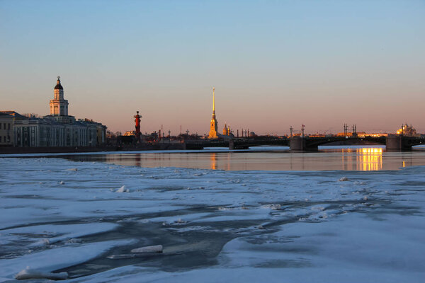 St. Petersburg, Russia - March 22, 2021. View of the Kunstkammer, Rostral Column, Peropavlovskaya Fortress and the Palace Bridge in the Neva riverbed with ice drift and reflexes in the water at sunset
