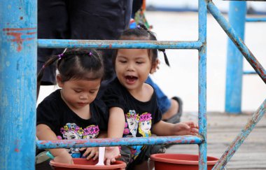 Pathumthani, Tayland - 6 Ağustos 2017: Duba üzerinde oturan ikiz kızın mutluluk anı ve Hong Pathummawas tapınağının önündeki nehir kenarında balık için Plastik kovada yem balığı hazırlamak.