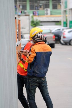 Pathumthani, Tayland - 6 Ağustos 2017: Turuncu ve mavi elbiseli iki inşaat işçisi yolun kenarındaki çinko duvarı elektrikli matkapla deliyorlar..
