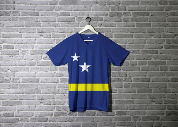 シャツにキュラソーの旗とレンガのパターンの壁紙と壁に掛け 中央と2つの白い星の少し下の水平黄色のストライプと青のフィールド — ストック写真
