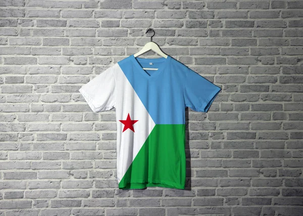 Gömlekte Cibuti bayrağı, duvarda da da tuğla desenli duvar kâğıdı asılı. Yatay açık mavi ve açık yeşil. Üzerinde kırmızı yıldız taşıyan beyaz bir üçgen var..