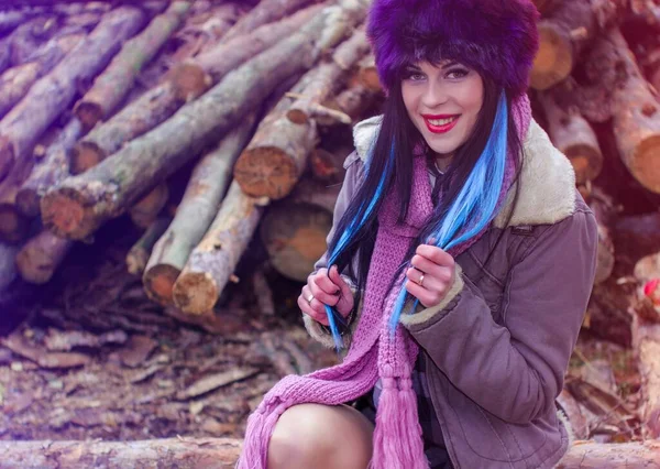 穿着假皮帽和夹克的现代美国风格的女人坐在森林里 一个有着长发和明亮假日的年轻女子在锯木厂化妆 — 图库照片