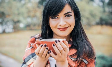 Genç, güzel bir kadın, klasik ekoseli bir elbiseyle gölün kenarında kahve içiyor. Sonbahar atmosferi, rahatlık kavramı