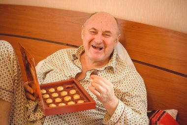 Yaşlı, mutlu, pijamalı bir adam sabahları yatağında çikolata tatlısı yer. Olgun erkeklerin psikolojisi, depresyon ve kötü ruh hallerinden kurtulmak.