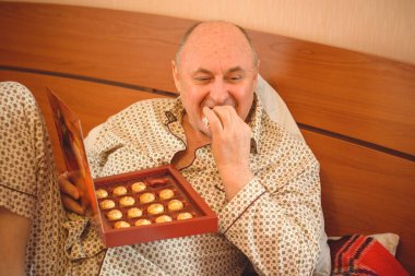 Yaşlı, mutlu, pijamalı bir adam sabahları yatağında çikolata tatlısı yer. Olgun erkeklerin psikolojisi, depresyon ve kötü ruh hallerinden kurtulmak.