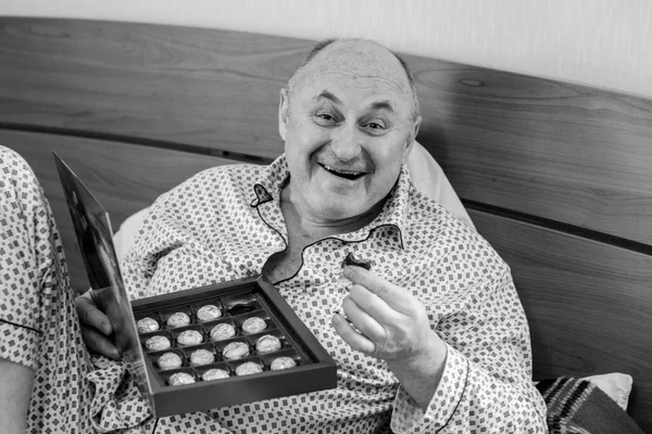 Yaşlı, mutlu, pijamalı bir adam sabahları yatağında çikolata tatlısı yer. Olgun erkeklerin psikolojisi, depresyon ve kötü ruh hallerinden kurtulmak.