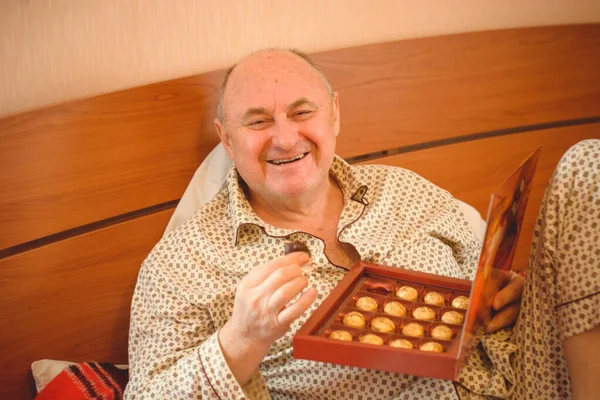 An elderly happy man in pajamas has chocolate sweets in bed in the morning. Psychology of mature men, getting rid of depression and bad mood