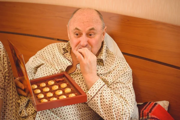 Yaşlı, mutlu, pijamalı bir adam sabahları yatağında çikolata tatlısı yer. Olgun erkeklerin psikolojisi, depresyon ve kötü ruh hallerinden kurtulmak.