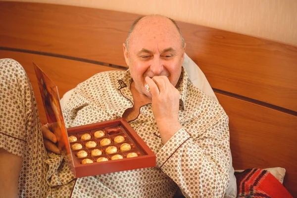 Yaşlı, mutlu, pijamalı bir adam sabahları yatağında çikolata tatlısı yer. Olgun erkeklerin psikolojisi, depresyon ve kötü ruh hallerinden kurtulmak.