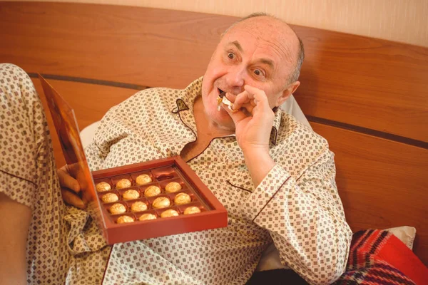 Yaşlı, mutlu, pijamalı bir adam sabahları yatağında çikolata tatlısı yer. Olgun erkeklerin psikolojisi, depresyon ve kötü ruh hallerinden kurtulmak.