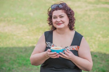 Orta yaşlı ve hayattan zevk alan iri yarı bir kadın. Aşırı kilolu olgun bir kadın, şık giyinmiş, olgun ve kilolu bir kadın ve bir fincan kahve.