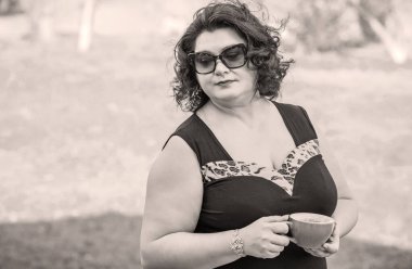 Orta yaşlı ve hayattan zevk alan iri yarı bir kadın. Aşırı kilolu olgun bir kadın, şık giyinmiş, olgun ve kilolu bir kadın ve bir fincan kahve.