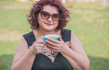 Orta yaşlı ve hayattan zevk alan iri yarı bir kadın. Aşırı kilolu olgun bir kadın, şık giyinmiş, olgun ve kilolu bir kadın ve bir fincan kahve.