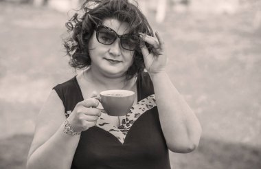 Orta yaşlı ve hayattan zevk alan iri yarı bir kadın. Aşırı kilolu olgun bir kadın, şık giyinmiş, olgun ve kilolu bir kadın ve bir fincan kahve.