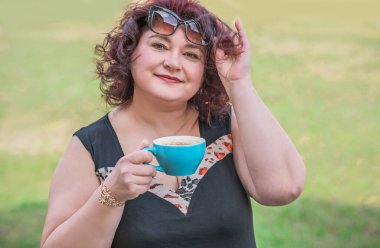 Orta yaşlı ve hayattan zevk alan iri yarı bir kadın. Aşırı kilolu olgun bir kadın, şık giyinmiş, olgun ve kilolu bir kadın ve bir fincan kahve.