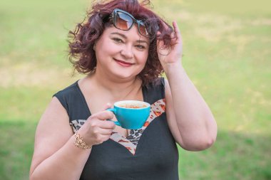 Orta yaşlı ve hayattan zevk alan iri yarı bir kadın. Aşırı kilolu olgun bir kadın, şık giyinmiş, olgun ve kilolu bir kadın ve bir fincan kahve.