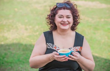 Orta yaşlı ve hayattan zevk alan iri yarı bir kadın. Aşırı kilolu olgun bir kadın, şık giyinmiş, olgun ve kilolu bir kadın ve bir fincan kahve.