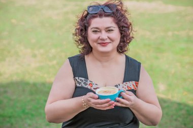 Orta yaşlı ve hayattan zevk alan iri yarı bir kadın. Aşırı kilolu olgun bir kadın, şık giyinmiş, olgun ve kilolu bir kadın ve bir fincan kahve.