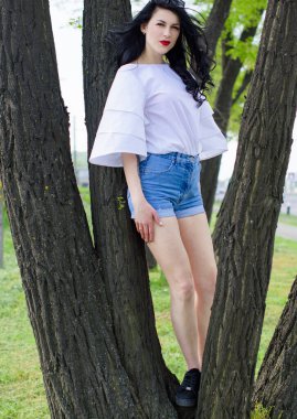 Şehir parkının ve bahçesinin arka planında siyah güneş gözlüğü takan güzel siyah saçlı genç kadın portresi. Beyaz romantik bluz ve yüksek bel kısa kot giyen bir kız..