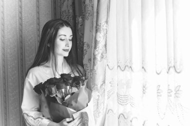 Bej rengi iç dairede bir buket kırmızı gülle romantik bir kadın. Konforlu evde ipek bornozlu bir bayan. Şefkat kavramı ve kadının hissi.