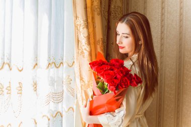 Bej rengi iç dairede bir buket kırmızı gülle romantik bir kadın. Konforlu evde ipek bornozlu bir bayan. Şefkat kavramı ve kadının hissi.