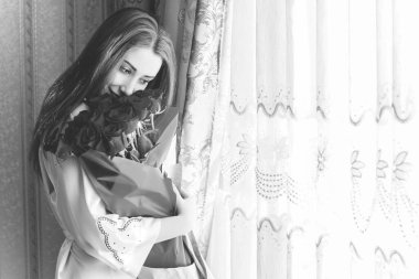 Bej rengi iç dairede bir buket kırmızı gülle romantik bir kadın. Konforlu evde ipek bornozlu bir bayan. Şefkat kavramı ve kadının hissi.