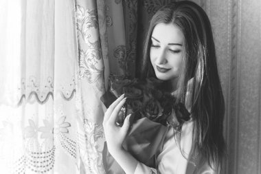 Bej rengi iç dairede bir buket kırmızı gülle romantik bir kadın. Konforlu evde ipek bornozlu bir bayan. Şefkat kavramı ve kadının hissi.