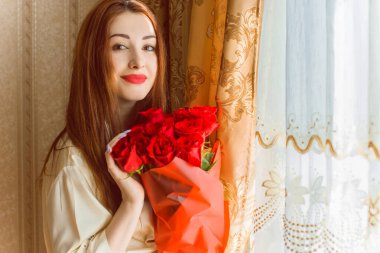 Bej rengi iç dairede bir buket kırmızı gülle romantik bir kadın. Konforlu evde ipek bornozlu bir bayan. Şefkat kavramı ve kadının hissi.