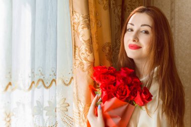 Bej rengi iç dairede bir buket kırmızı gülle romantik bir kadın. Konforlu evde ipek bornozlu bir bayan. Şefkat kavramı ve kadının hissi.