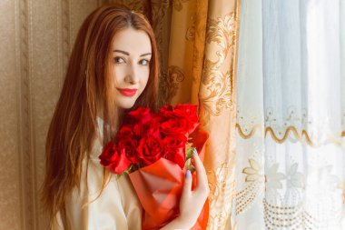 Bej rengi iç dairede bir buket kırmızı gülle romantik bir kadın. Konforlu evde ipek bornozlu bir bayan. Şefkat kavramı ve kadının hissi.