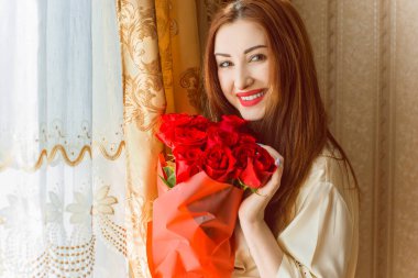 Bej rengi iç dairede bir buket kırmızı gülle romantik bir kadın. Konforlu evde ipek bornozlu bir bayan. Şefkat kavramı ve kadının hissi.