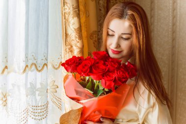 Bej rengi iç dairede bir buket kırmızı gülle romantik bir kadın. Konforlu evde ipek bornozlu bir bayan. Şefkat kavramı ve kadının hissi.