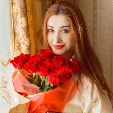 Bej rengi iç dairede bir buket kırmızı gülle romantik bir kadın. Konforlu evde ipek bornozlu bir bayan. Şefkat kavramı ve kadının hissi.