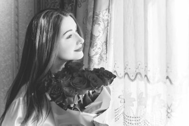 Bej rengi iç dairede bir buket kırmızı gülle romantik bir kadın. Konforlu evde ipek bornozlu bir bayan. Şefkat kavramı ve kadının hissi.