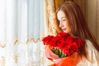 Bej rengi iç dairede bir buket kırmızı gülle romantik bir kadın. Konforlu evde ipek bornozlu bir bayan. Şefkat kavramı ve kadının hissi.