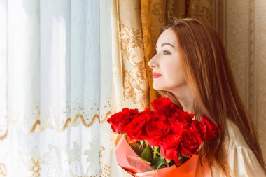 Bej rengi iç dairede bir buket kırmızı gülle romantik bir kadın. Konforlu evde ipek bornozlu bir bayan. Şefkat kavramı ve kadının hissi.