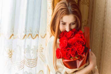 Bej rengi iç dairede bir buket kırmızı gülle romantik bir kadın. Konforlu evde ipek bornozlu bir bayan. Şefkat kavramı ve kadının hissi.